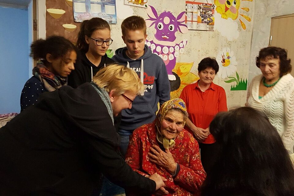 Eine Frau beugt sich zu einer älteren Frau herunter und nimmt ihre Hand. Im Hintergrund stehen junge Leute