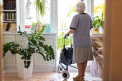 Die meisten Menschen möchten auch im Alter zu Hause leben. Eine alte Frau mit Rollator schaut aus dem Fenster ihrer Wohnung