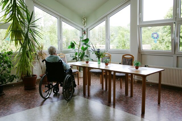 Anke Thomass/stock.adobe.com Eine alte Frau sitzt im Rollstuhl allein an einem Tisch in einem Pflegeheim